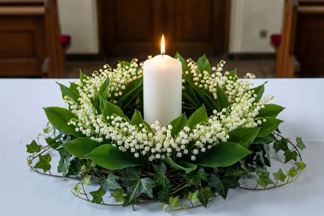 Een brandende witte doopkaars staat in een krans van meiklokjes en afhangende klimop op een wit tafelkleed in een kerk.
