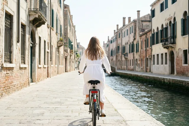 Een meisje met lang haar en een wit gewaad fietst van de camera weg langs een kanaal met oude gebouwen, boogramen en luiken in helder daglicht.