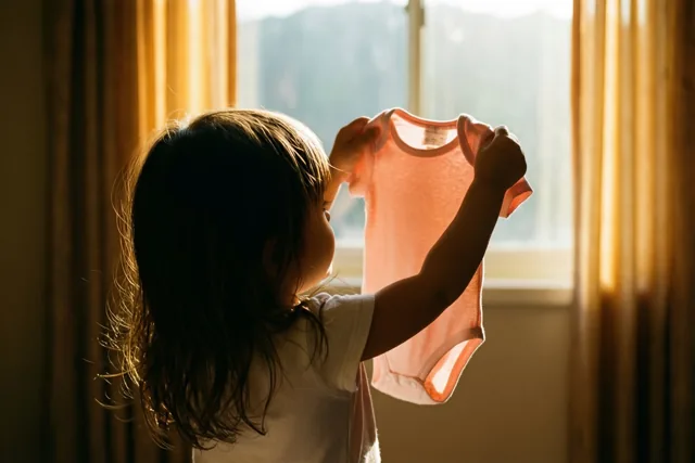 Een meisje met donker haar staat met haar rug naar de camera en houdt een roze babypakje naar het heldere raamlicht, terwijl gouden zonlicht door de gordijnen valt.