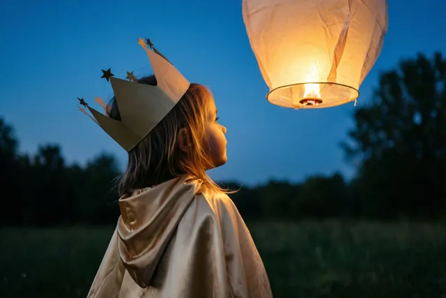 Een jong meisje met een papieren kroon en een gouden cape kijkt in een veld met bomen naar een lichtgevende hemellantaarn tegen de schemerlucht.