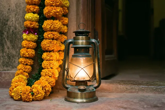 Een brandende messing lantaarn staat op een stenen vloer naast een bloemenslinger tegen de muur van een zwak verlichte deuropening, in gouden, oranje en bronzen tinten.