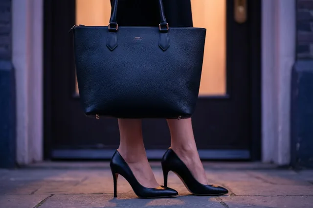 Een persoon met zwarte schoenen met hoge hakken houdt een zwarte leren draagtas vast bij een voordeur, in zacht en warm licht.