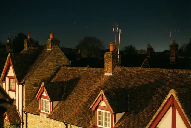 Bij een schoorsteen boven warm verlichte trapgevels van stenen gebouwen en vakwerkhuizen tekenen een rode mijter en gouden staf zich af tegen een donkere hemel met zwakke sterren.