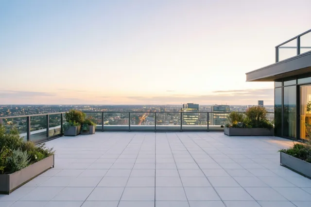 Een ruim dakterras met lichtgrijze tegels, glazen balustrades en planten in potten kijkt uit over oplichtende stadslichten onder een lucht die van lichtblauw naar warm oranje verloopt.