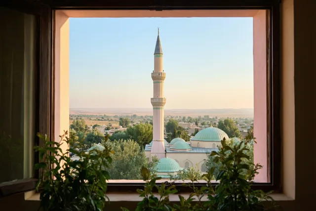 Door een raam is een moskee met een slanke minaret te zien onder een heldere lichtblauwe ochtendlucht, met groen op de voorgrond.