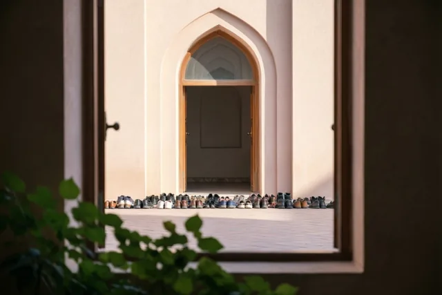 Door een raam is een boogvormige ingang van de moskee te zien met netjes opgestelde schoenen in warm ochtendzonlicht en groen dat een deel van de voorgrond bedekt.