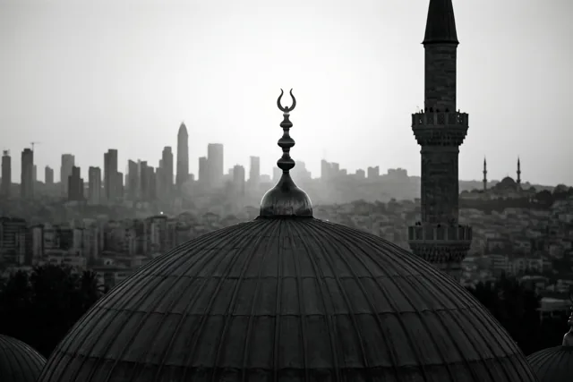 Een zwart-witfoto toont het silhouet van een moskeekoepel met een maansikkel, een minaret rechts en een wazig stadsgezicht op de achtergrond.