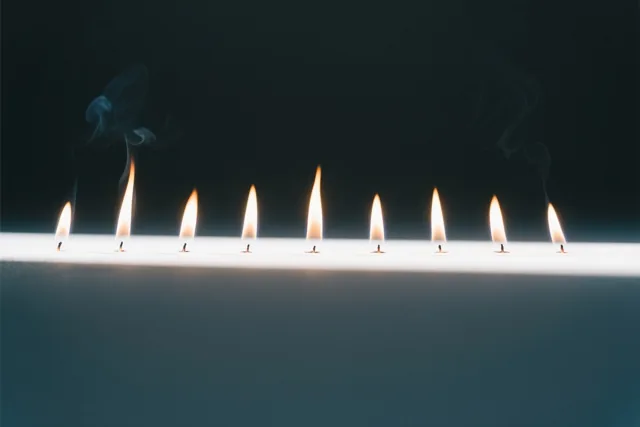 Negen kleine vlammen branden in een rechte lijn tegen een donkere achtergrond, met warm oranje licht en koele blauwgrijze tinten.