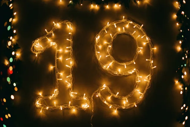 Warme lichtjes vormen 19 op een donkere muur, met onscherpe feestverlichting op de achtergrond.