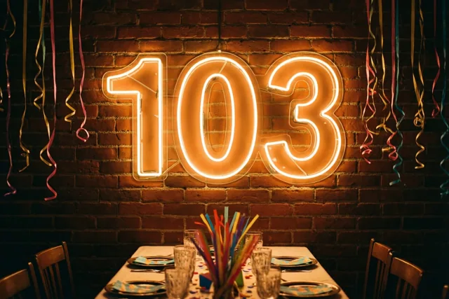 Een neonbord met 103 licht warm op aan een bakstenen muur boven een gedekte feesttafel, met kleurrijke slingers aan het plafond.