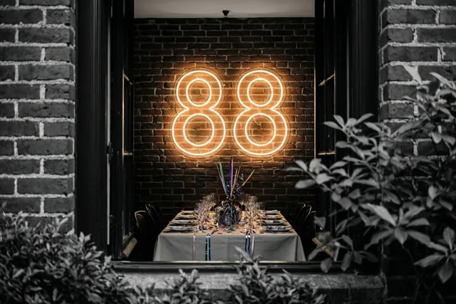 Een warm neongetal 88 hangt boven een feestelijk gedekte tafel aan een bakstenen muur, gezien door een open deuropening met gebladerte eromheen.