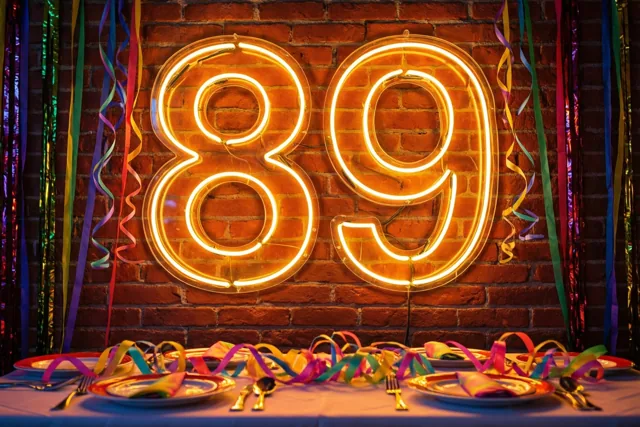 Een groot warm oplichtend neongetal 89 hangt aan een rode bakstenen muur boven een feestelijk gedekte tafel met kleurrijke slingers en borden.