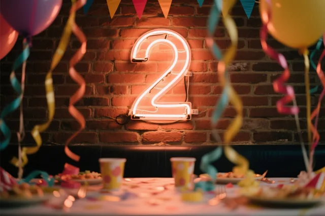 Een lichtgevende neon-2 hangt aan een bakstenen muur boven een feesttafel met kleurrijke slingers en ballonnen.