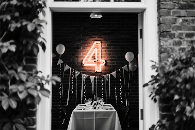 Een warm verlicht neoncijfer 4 hangt aan een donkere bakstenen muur boven een gedekte feesttafel met slingers en ballonnen, omlijst door groen gebladerte.