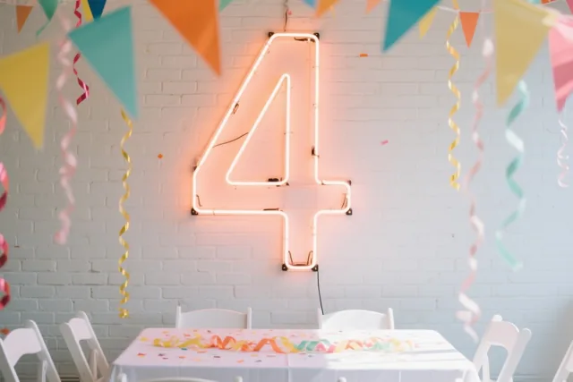 Een groot neoncijfer 4 hangt aan een witte bakstenen muur boven een tafel met een wit tafelkleed, kleurrijke slingers, vlaggetjes en spiraalslingers.
