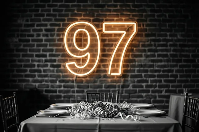 Een feesttafel met slingers staat onder een warm oranje neongetal 97 aan een bakstenen muur, tegen een donkere achtergrond.