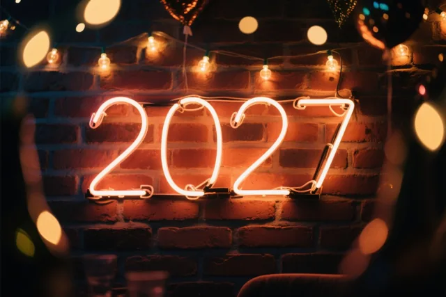 Een neonbord met het jaartal 2027 hangt aan een warm verlichte bakstenen muur, met lichtslingers en onscherpe lichtjes eromheen.