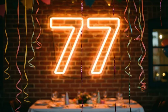 Een warm gloeiend neongetal 77 hangt aan een bakstenen muur boven een versierde feesttafel met kleurrijke slingers.