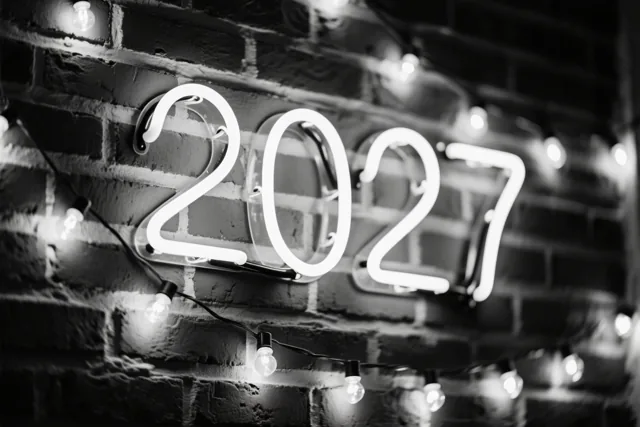 Een zwart-witfoto toont een lichtgevend neonbord met 2027 op een bakstenen muur, met lichtslingers rond het bord.