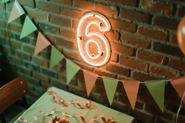 Een lichtgevend neoncijfer 6 hangt aan een rode bakstenen muur boven een feesttafel met groene en perzikkleurige vlaggetjes en slingers.
