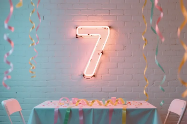 Een lichtgevend neoncijfer 7 hangt aan een witte bakstenen muur boven een feesttafel met slingers en confetti, in roze en blauw licht.