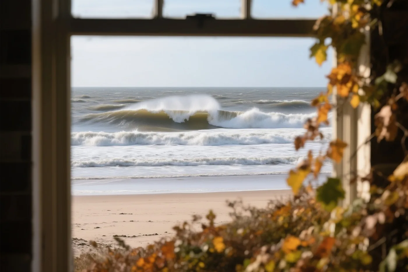 Door een raam met herfstbladeren op de voorgrond zijn hoge golven op een leeg Noordzeestrand te zien in blauwe en grijze tinten.