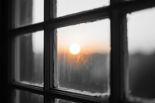 Door een natte ruit van een raam met meerdere vakken schijnt een heldere opkomende zon, met oranje en witte tinten tegen een monochrome achtergrond.