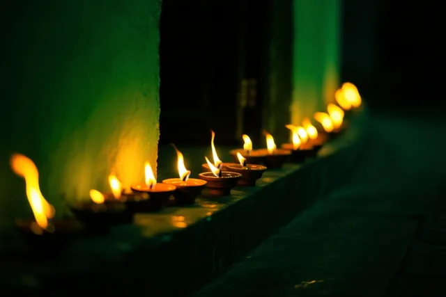 Een rij kleine olielampjes met flakkerende vlammen verlicht een donkere drempel met warm geel licht tegen een groene muur.