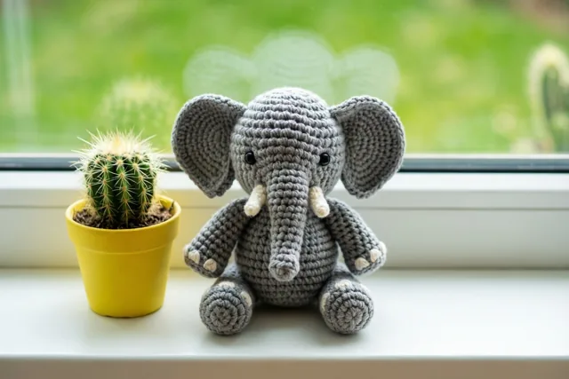 Een klein gehaakt grijs olifantje zit op een witte vensterbank naast een cactus in een gele pot, in daglicht met onscherp groen buiten.