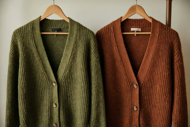 Een olijfgroen en een roestbruin gebreid vest hangen naast elkaar op houten kleerhangers tegen een effen muur in zacht licht.