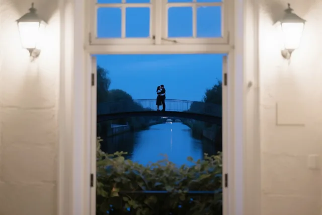 Door een open deuropening met brandende wandlampen is in de verte een koppel te zien dat elkaar omhelst op een voetbrug over een kanaal in de schemering.