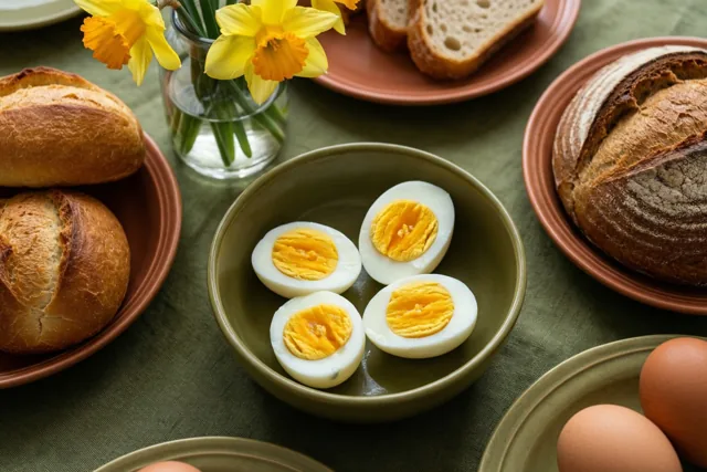 Een kom met gehalveerde gekookte eieren, brood op terracotta borden en een vaas gele narcissen staan op een groen tafelkleed.