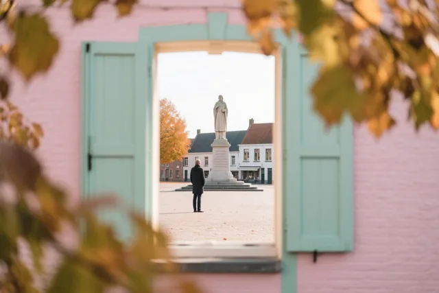 Door een open raam met roze muren en herfstbladeren is een persoon bij een oorlogsmonument op een Vlaams dorpsplein te zien.