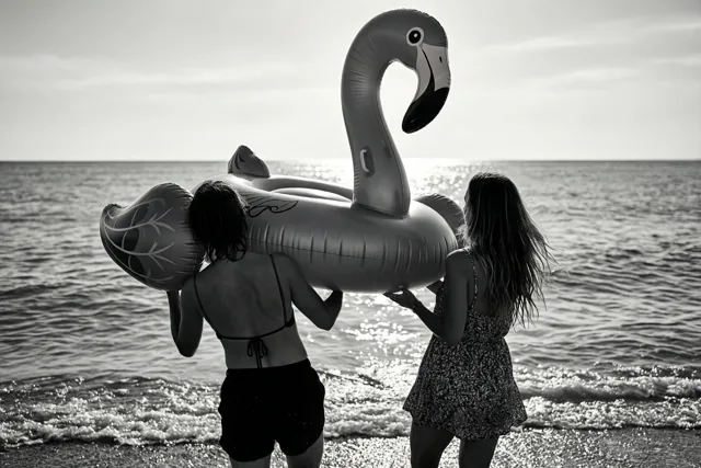Twee mensen dragen met hun rug naar de camera een grote opblaasflamingo naar een kalme zee op een zwart-witfoto met fel zonlicht op het water.