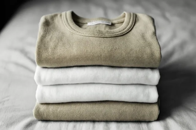 Een nette stapel opgevouwen babykleertjes in beige en wit ligt op een zacht oppervlak met structuur bij natuurlijk licht.