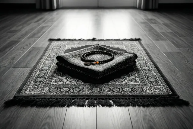 Een opgevouwen gebedskleed met gebedskralen ligt in het ochtendlicht op een houten vloer, in een vrijwel monochroom beeld met warme tinten in de kralen.
