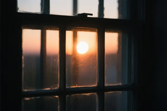 Vanuit een schemerige kamer zie je door een raam met meerdere ruitjes een stralende opkomende zon in warm oranje en geel licht.