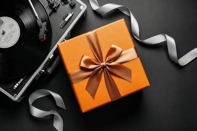 Een oranje geschenkdoos met een bruine strik staat naast een zwarte platenspeler op een donkere ondergrond.