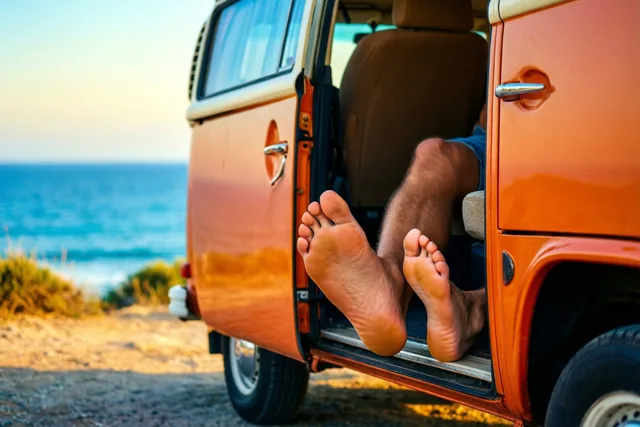 Een oude oranje camper staat op een onverhard kustpad met blote voeten uit de open zijdeur en een blauwe zee en lucht in het warme zonlicht.
