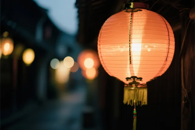 Een brandende oranje papieren lampion is van dichtbij te zien in een donkere straat, met onscherpe lichtjes en koele blauwe tinten op de achtergrond.