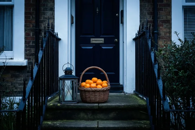 Een mand mandarijnen en een lantaarn staan op de stenen treden voor de deur van een rijhuis, met feloranje fruit tegen koele blauwe en zwarte tinten.