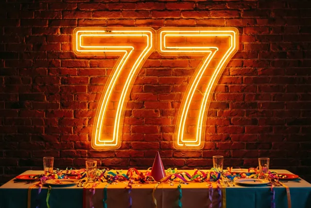 Oranje neonlicht vormt 77 op een rode bakstenen muur boven een tafel met een blauw tafellaken, kleurrijke slingers en feestversiering.