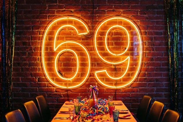 Een groot oranje neongetal 69 hangt aan een rode bakstenen muur boven een feesttafel met kleurrijke slingers en glazen.