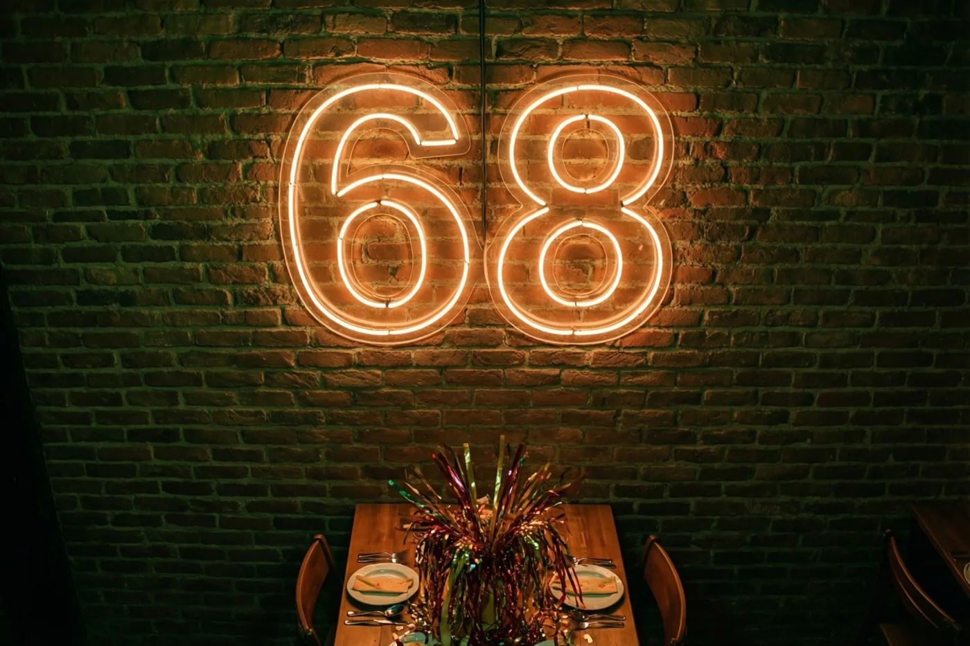 Een oranje neongetal 68 hangt op een donkere bakstenen muur boven een houten feesttafel met borden, bestek en kleurrijke slingers in het midden.