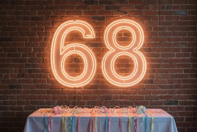 Een groot oranje neongetal 68 hangt aan een rode bakstenen muur boven een feesttafel met een wit kleed, kleurrijke slingers en confetti.