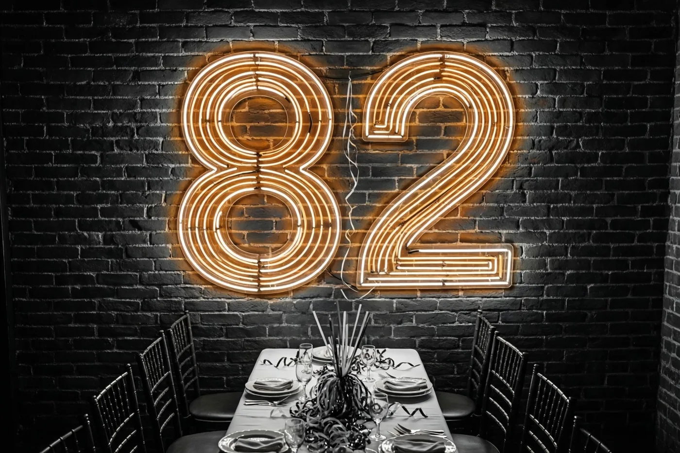 Oranje neonlicht vormt 82 op een donkere bakstenen muur boven een formeel gedekte feesttafel in zwart-wittinten.