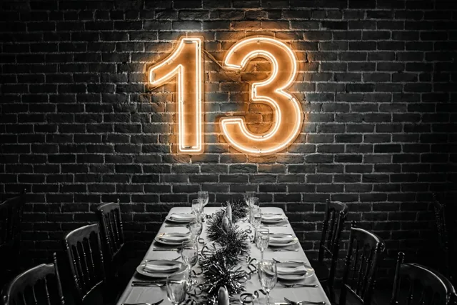 Een oranje neongetal 13 hangt aan een donkere bakstenen muur boven een lange feesttafel met zwart-wit servies en zilveren glinsterende versiering.