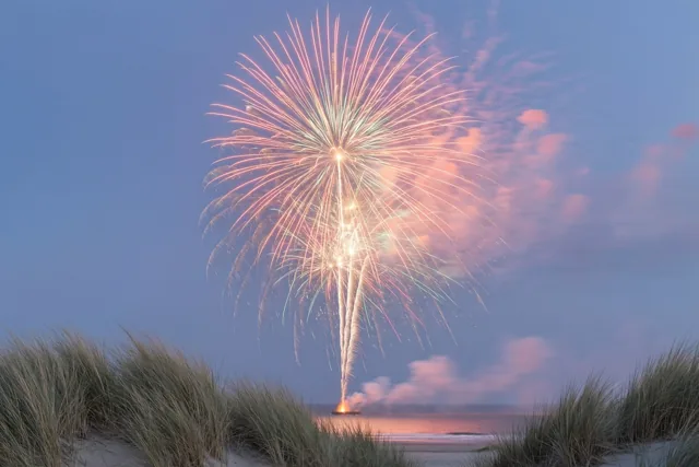 Een grote vuurwerkexplosie in oranje en roze verlicht de blauwe schemerlucht boven de zee, met zandduinen en strandgras op de voorgrond.