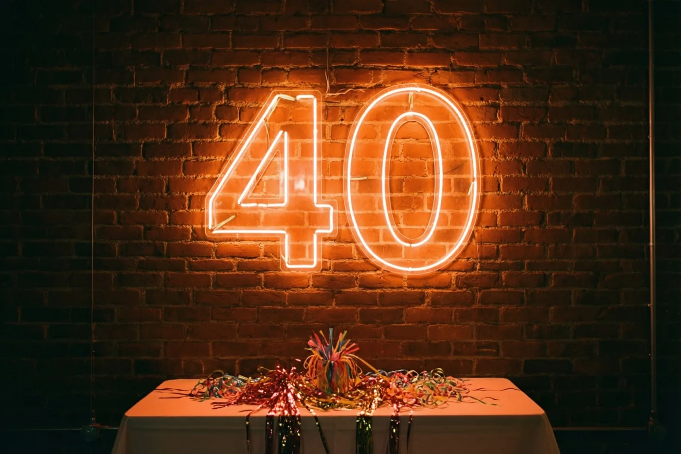 Een oranje neongetal 40 hangt aan een donkere bakstenen muur boven een tafel met een wit kleed en kleurrijke serpentines.