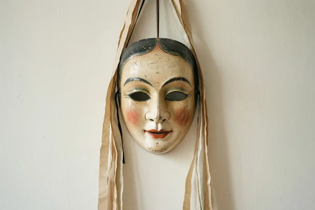 Een oud masker van papier-maché met geschilderde gelaatstrekken, gebarsten verf en vervaagde kleuren hangt aan beige papieren slingers tegen een effen muur.